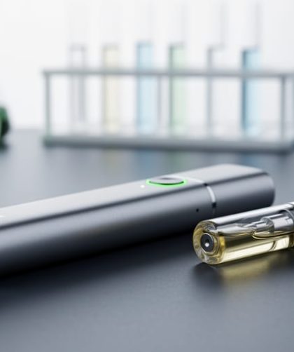 Close-up of a vape device and oil cartridge on a dark surface with blurred laboratory-like equipment in the background.